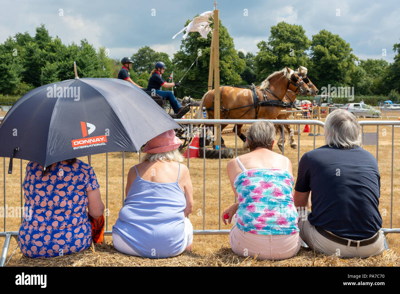 People sitting on hay bales watching a heavy horse event at a country fair in Hampshire, UK, in hot summer sunshine. Two people sit under a large sun umbrella for shade. Stock Photo