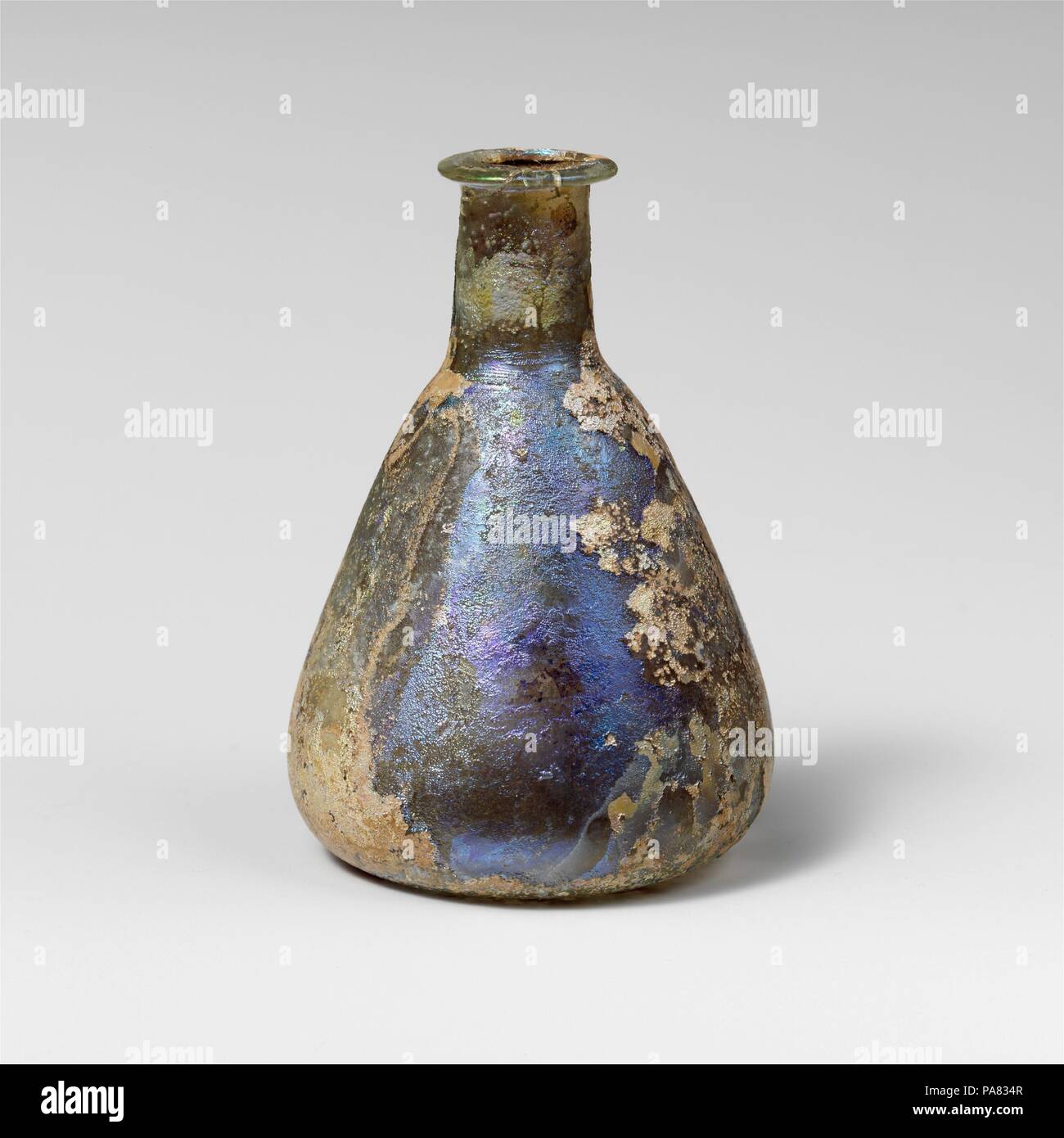 Glass perfume bottle. Culture: Roman. Dimensions: Overall: 3 3/16in. (8.1cm)  Diam.: 2 3/16 x 13/16 in. (5.6 x 2.1 cm). Date: 1st century A.D..  Colorless with pale blue green tinge.  Rim folded out, over, and in, flattened on top; short, cylindrical neck, with tooling marks around base; conical body, rounded at base; concave bottom.  Complete, but broken and repaired on one side of rim; many pinprick bubbles; deep pitting, dulling, and iridescent weathering.  Contains large black (burnt ?) solid object. Museum: Metropolitan Museum of Art, New York, USA. Stock Photo