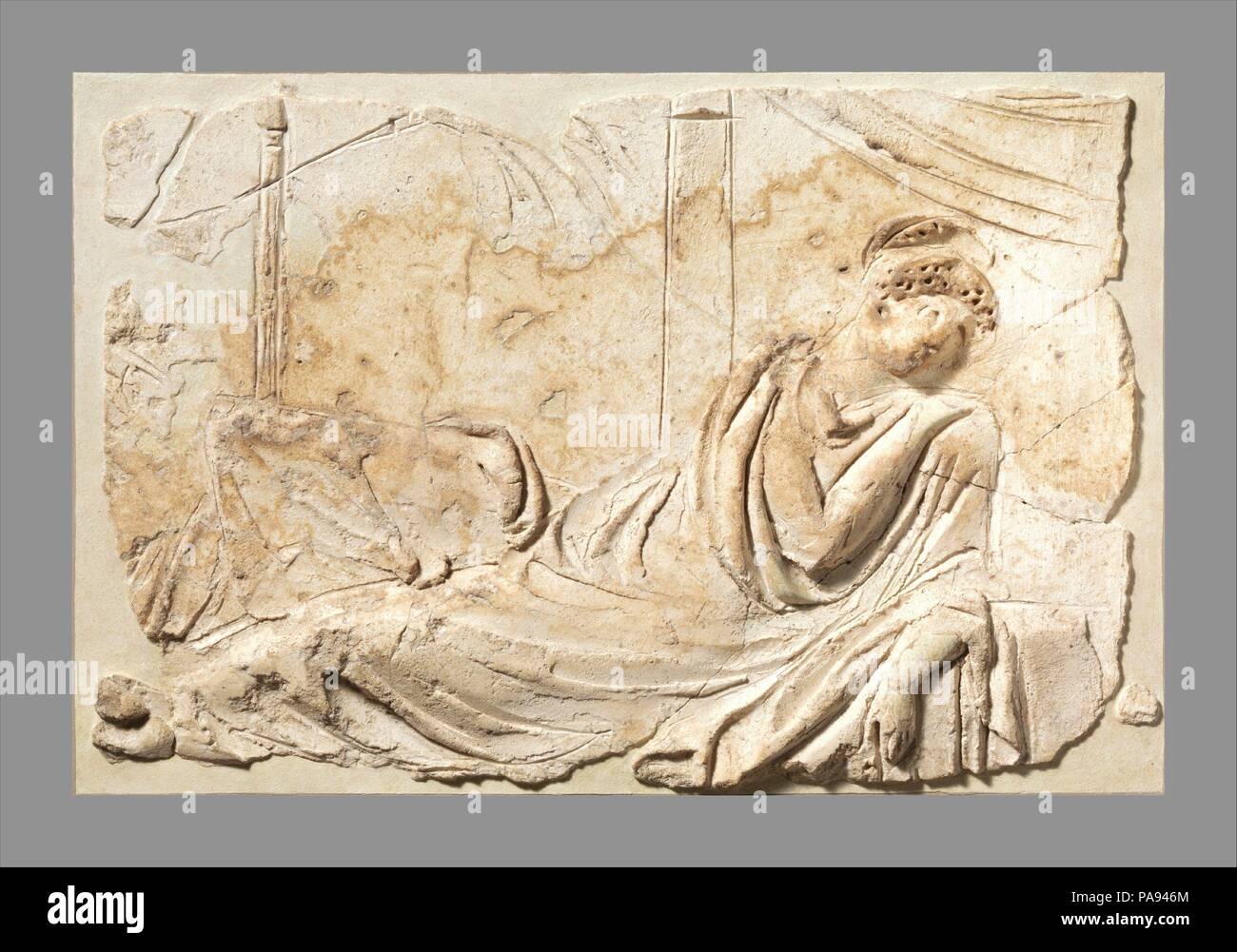 Stucco relief panel. Culture: Roman. Dimensions: Overall: 9 1/2 x 14 3/8 x 2 1/4 in. (24.1 x 36.5 x 5.7 cm). Date: 2nd half of 1st century A.D..  Draped in a heavy cloak and with an elaborate hairdo, the woman reclines with one knee raised. Her left arm rests against a low support. A pillar from which a swag of drapery hangs is visible just behind her right shoulder and another slender, fluted column stands to the left. A long, thin palm-like branch sketched lightly in the background at the upper left must have been held by another figure, who was approaching the resting woman. Given the Diony Stock Photo