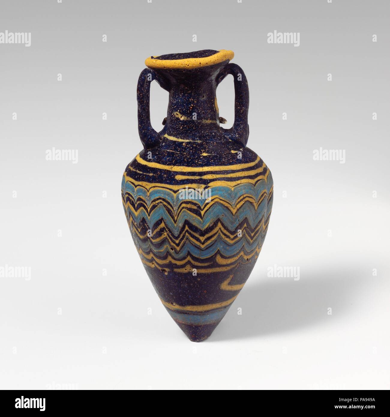 Glass amphoriskos (perfume bottle). Culture: Greek, Eastern Mediterranean. Dimensions: H. 3 9/16 in. (9 cm). Date: late 6th-5th century B.C..  Translucent dark cobalt blue, with handles in same color; trails in opaque yellow and opaque turquoise blue.  Broad inward-sloping rim-disk; cylindrical neck, expanding slightly downwards; broad, angular shoulder; tall conical body, tapering downwards to pointed bottom; two vertical strap handles applied to top of shoulder, drawn up, and pressed onto underside of rim-disk.  One yellow trail attached at edge of rim-disk; another yellow trail applied to n Stock Photo