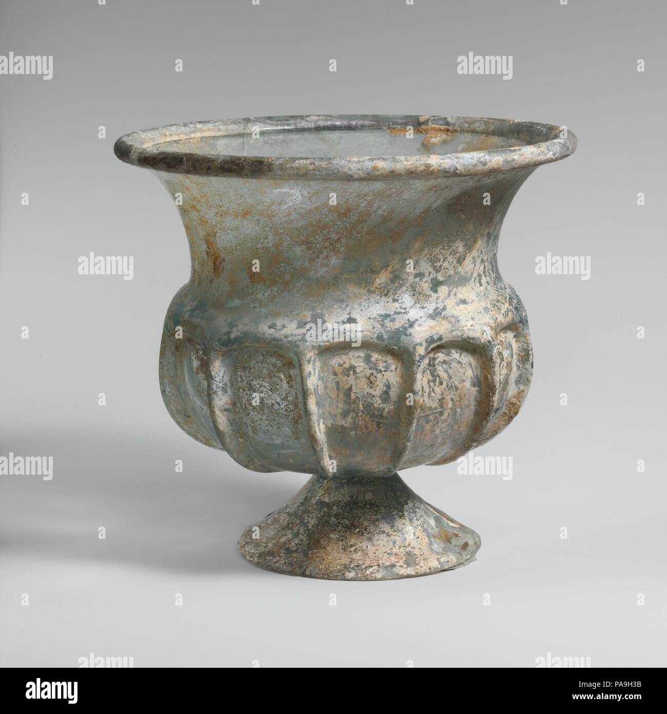 Glass cup with splayed foot. Culture: Roman. Dimensions: H. 5 3/16 in. (13.2 cm); diameter  5 9/16 in. (14.1 cm). Date: 2nd half of 1st century A.D..  Translucent blue green.  Outsplayed tubular rim, folded over and in; tall curving neck, tapering downwards; thick projecting shoulder; squat, globular body; applied conical foot, formed from a separate gather, with edge cracked-off and ground flat; small, circular, flat bottom.  Eleven vertical ribs in high relief tooled out from shoulder down side of body.  Rim and body cracked with one hole in side; part of foot broken and missing; few bubbles Stock Photo