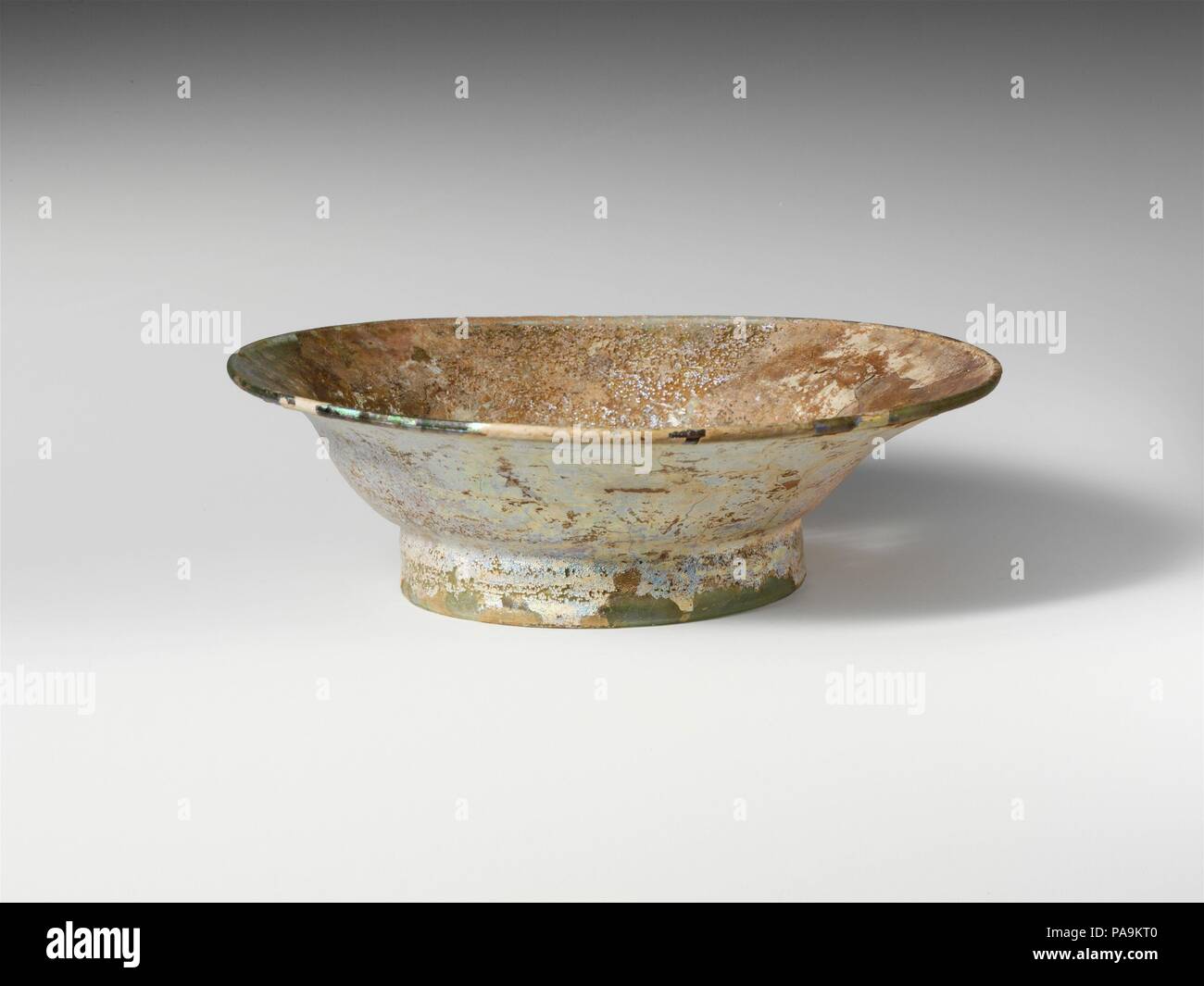 Glass dish. Culture: Roman. Dimensions: H.: 2 5/16 in. (5.9 cm)  Diam.: 7 3/16 x 3 3/4 in. (18.3 x 9.5 cm). Date: 4th century A.D..  Translucent pale blue green.  Thickened, rounded rim; side curves down and in, with angular profile; tall foot ring, made by folding, slightly splayed and with tubular, inward-bulging edge; uneven bottom with thick dome at center and prominent pontil scar.   Intact; many pinprick bubbles; heavily weathered, with pitting, some dulling, and brilliant iridescence. Museum: Metropolitan Museum of Art, New York, USA. Stock Photo