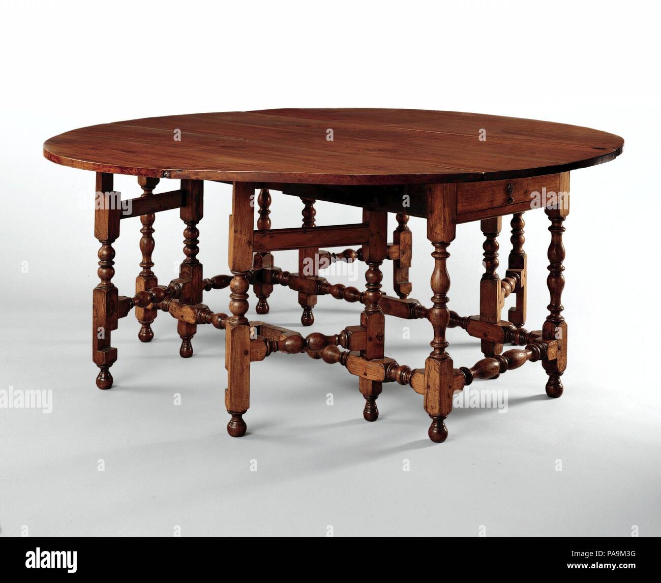 Oval table with falling leaves. Culture: American. Dimensions: 29 3/4 x 74 1/2 x 71 in. (75.6 x 189.2 x 180.3 cm). Date: 1690-1720.  This spectacular walnut table is as fine as any made in colonial America. Its multiple turned legs and gate-like supports create a virtual forest of baluster turnings. In stark contrast to the rectilinearity of seventeenth-century tables, the gentle elliptical curve of its top invited conviviality and conversation among those seated around it. The narrow width of leather and cane chairs of this period were ideal for use among the multiple legs on tables like thes Stock Photo