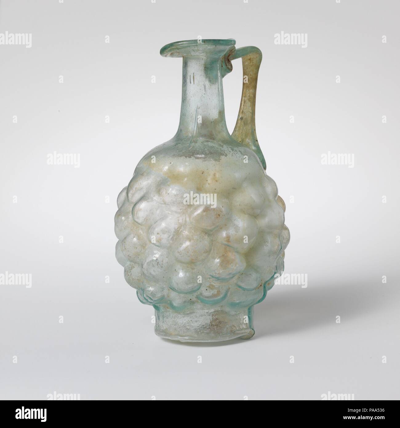 Glass jug in the form of a pine cone. Culture: Roman. Dimensions: H.: 4 3/8 in. (11.1 cm). Date: 2nd half of the 1st century A.D..  Translucent blue green; handle in same color.  Misshapen thick, everted rim with fire-rounded lip, half turned downwards, half flaring upwards; cylindrical neck expanding downwards; plain, convex shoulder; globular body; tall circular base, with slightly concave, uneven bottom; strap handle attached to shoulder and top of body, drawn up and out, then turned in at an acute angle, and trailed on to underside of rim and top of neck. One continuous mold seam around bo Stock Photo