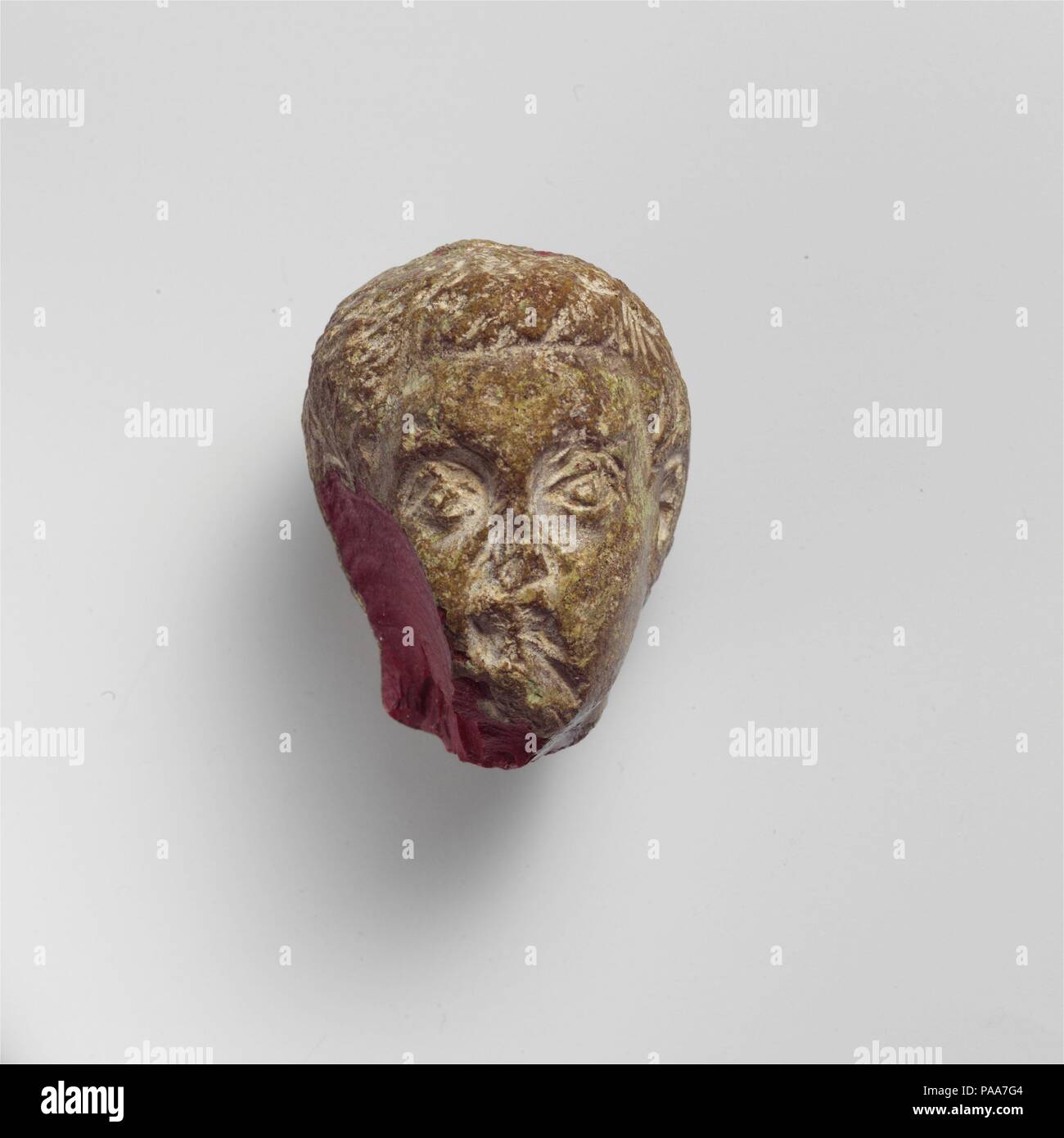 Glass portrait head. Culture: Roman. Dimensions: Height: 1 15/16 in. (5 cm). Date: 1st half of 4th century A.D..  Opaque deep red.  Solid block.  Carved in the round: male head with short wavy hair and fringe across forehead, arched brows, large eyes, and clean shaven face.  Broken, with large unweathered chip in proper right side of face and side of head, and weathered chips to nose, mouth, and chin; green copper weathering surface.  This small portrait head may represent a prince from the family of Constantine I. Museum: Metropolitan Museum of Art, New York, USA. Stock Photo