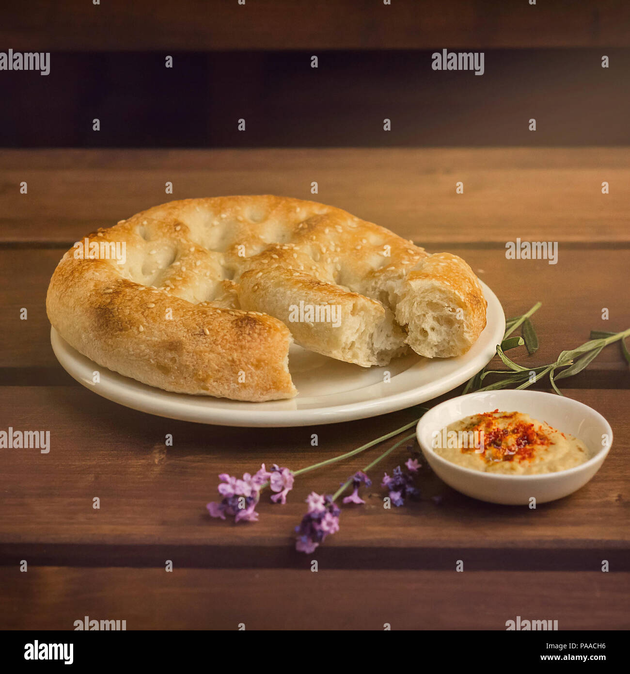 Round fresh bread, sprinkled with sesame seeds, lies on a plate. Next to a small plate with sauce and a branch of lavender Stock Photo