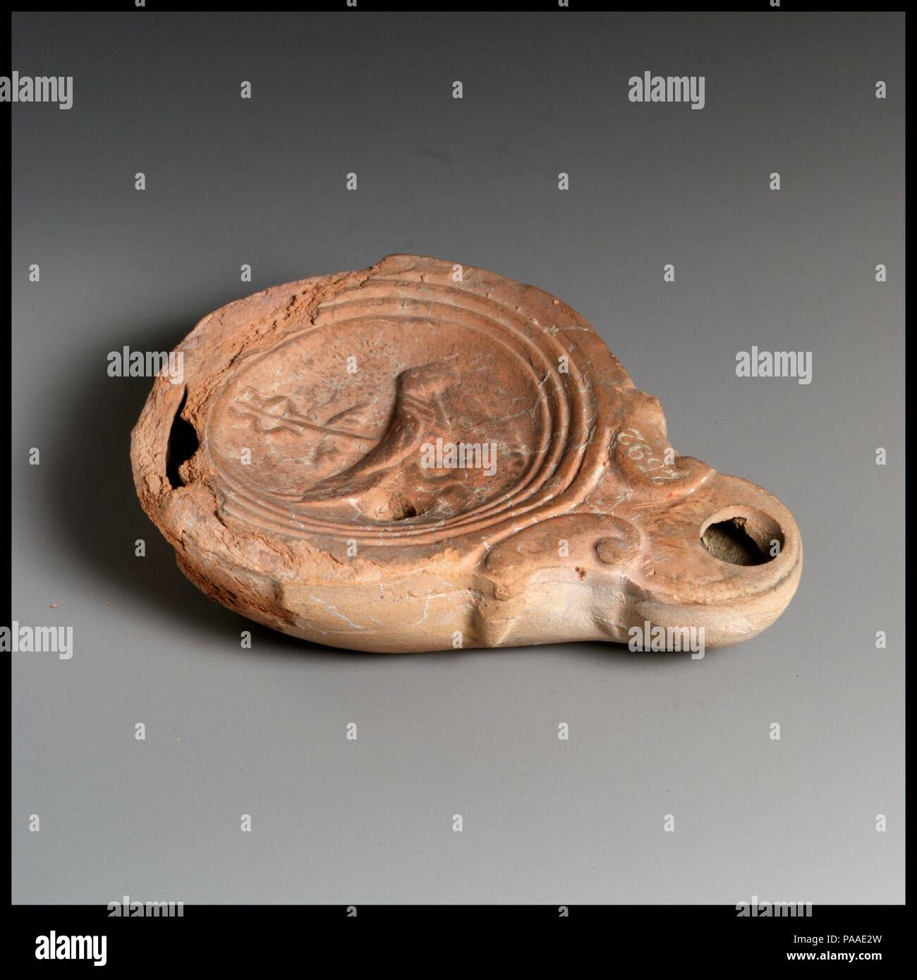 Terracotta oil lamp. Culture: Roman. Dimensions: Overall: 1 x 3 1/2 in. (2.5 x 8.9 cm). Date: 1st century B.C.-1st century A.D..  Loeschcke Type 4. Mold-made. Discus: a bird (an eagle or parrot?), facing right, holding a ribbon in its curved beak, standing in front of a winged caduceus, slanting obliquely to left; filling holelling hole at lower left behind the bird's tail, and a band of lines and grooves toward edge. Small volutes flanking nozzle, with off-center wick hole. Incised base ring, and concave base.  Hole and extensive chipping to shoulder and top of body at back. Museum: Metropoli Stock Photo