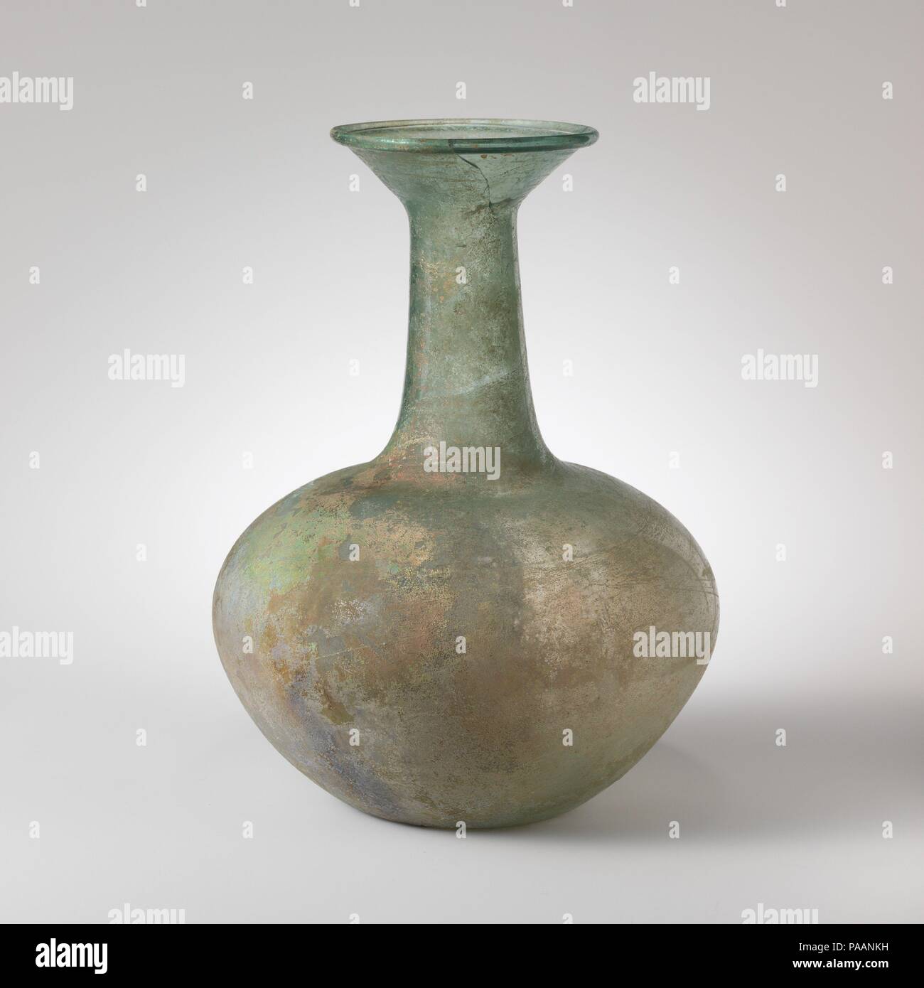 Glass bottle. Culture: Roman. Dimensions: H.: 9 7/16 in. (24 cm). Date: 4th-5th century A.D..  Translucent blue green.  Everted tubular rim, folded over and in; funnel-shaped mouth; cylindrical neck, expanding downwards; broad but squat body with maximum diameter above mid-point; concave bottom.  Broken and repaired on one side of rim and mouth; pinprick bubbles and blowing striations; dulling, pitting, and iridescent weathering. Museum: Metropolitan Museum of Art, New York, USA. Stock Photo