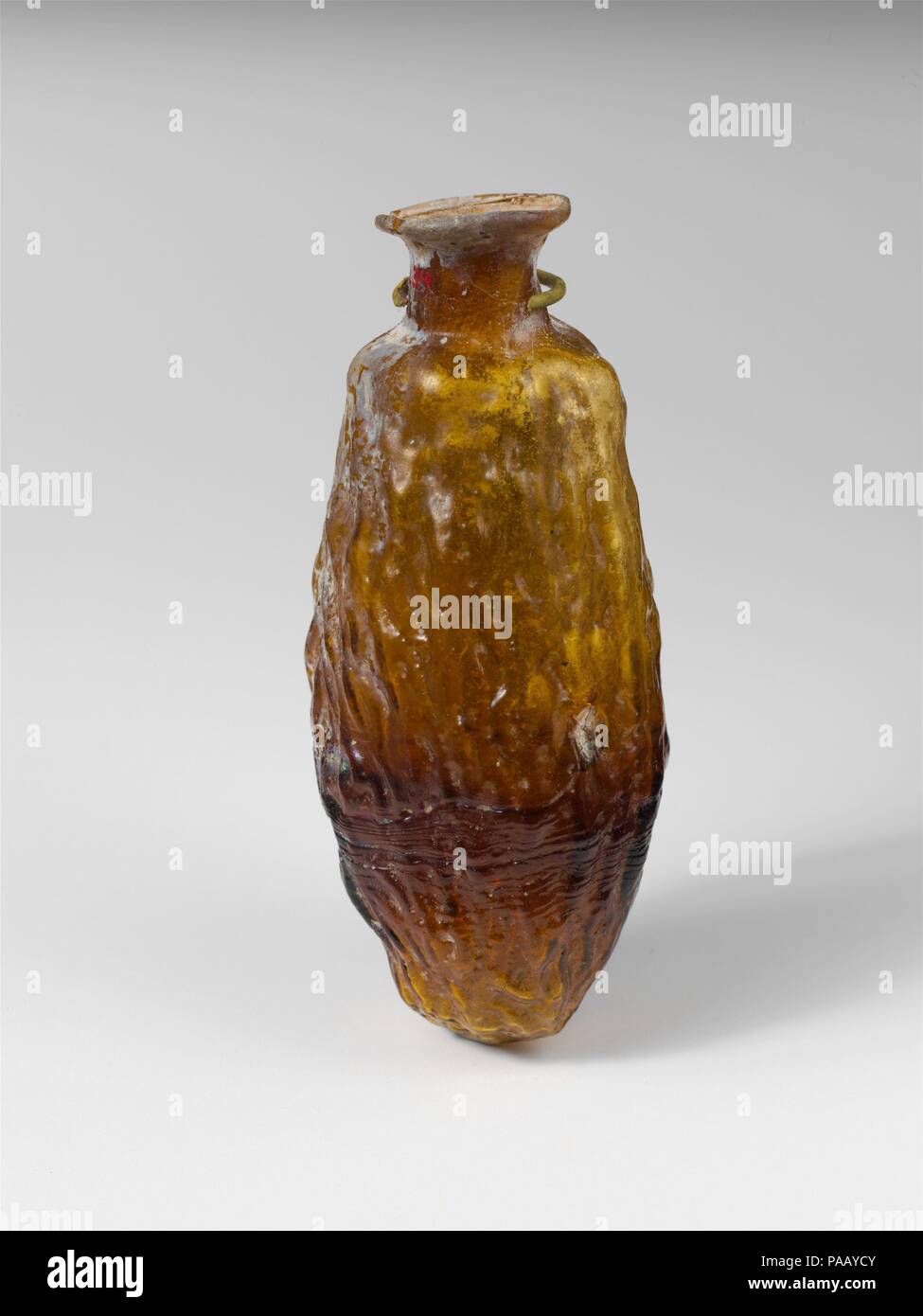 Glass bottle shaped like a date. Culture: Roman. Dimensions: H.: 2 7/8 in. (7.3 cm)  Diam.: 5/8 x 1 3/16 in. (1.6 x 3 cm). Date: mid-1st to early 2nd century A.D..  Translucent yellowish brown.  Thin, everted rim folded round and in; flaring neck with concave sides; elongated body; round bottom. One continuous mold seam around body.  Body molded into the shape of a wrinckled date, with pattern of short wavy ridges and hollows.  Intact, except for small chip and crack in rim; some bubbles and blowing striations; slight dulling, iridescence, and whitish weathering. Museum: Metropolitan Museum of Stock Photo