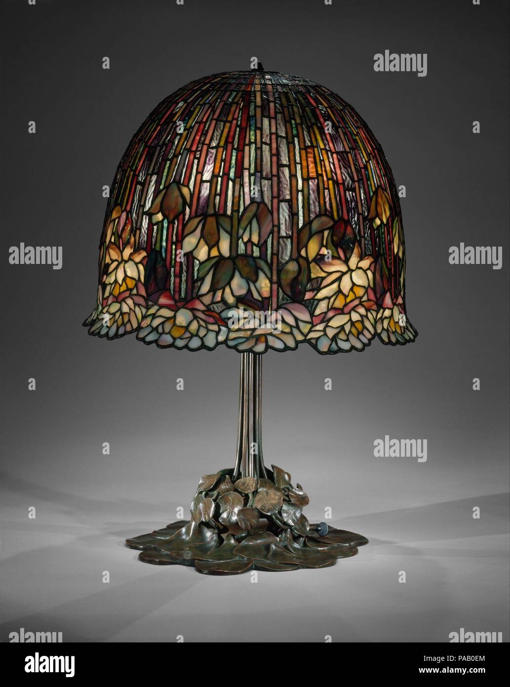 Lamp. Culture: American. Designer: Designed by Louis Comfort Tiffany (American, New York 1848-1933 New York). Dimensions: Base: H. 26 1/2 in. (67.3 cm); Diam. 14 5/8 in. (37.1 cm)  Shade: H. 14 5/8 in. (37.1 cm). Maker: Tiffany Studios (1902-32). Date: 1904-15.  This water-lily table lamp is one of Tiffany's most successfully executed designs for his firm's well-known leaded-glass products. The overall organic character of the lamp is emphasized throughout the piece in its details. The bronze support replicates broad, flat lily pads clustered around a base, out of which rise attenuated climbin Stock Photo