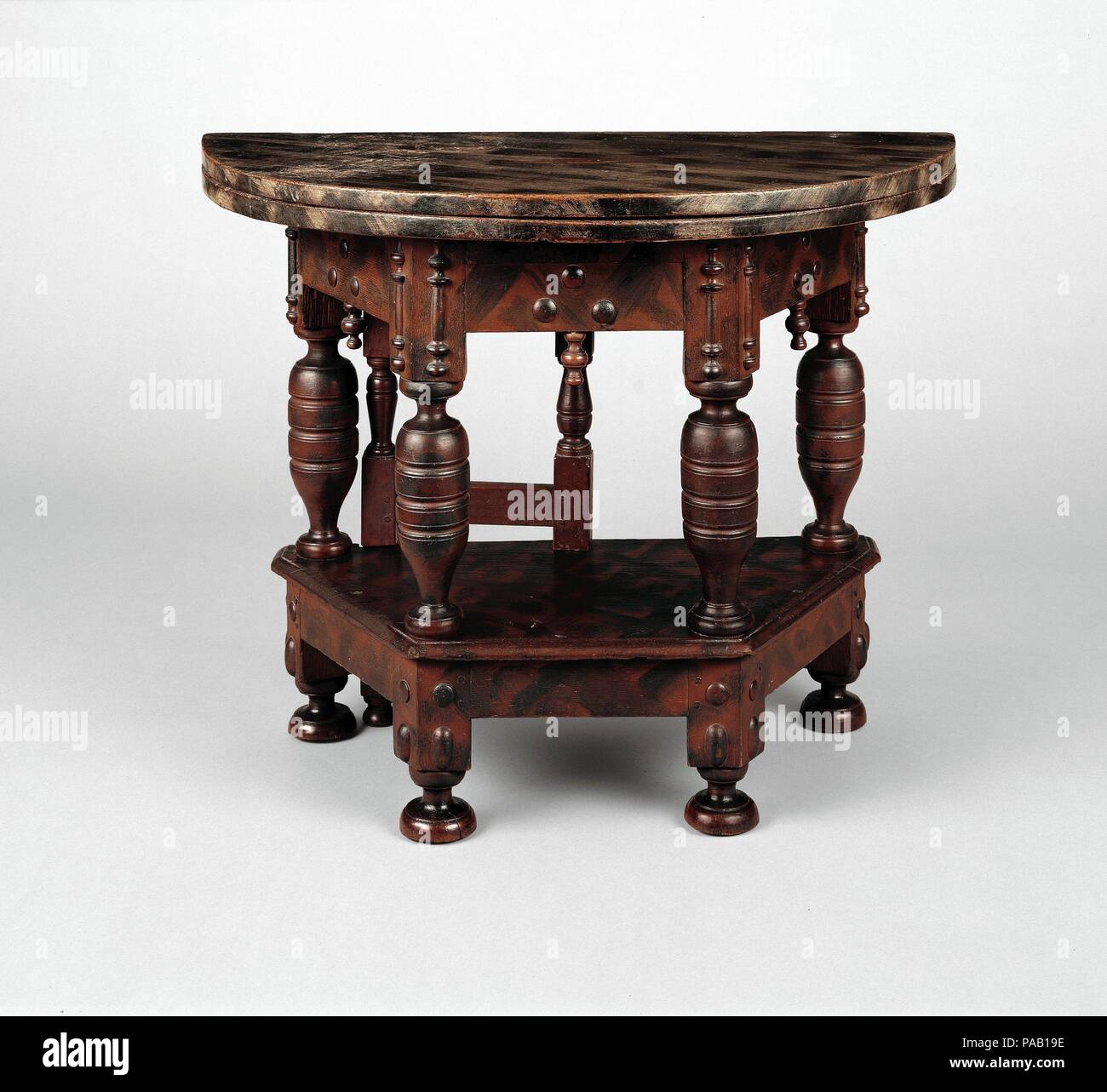 Folding table. Culture: American. Dimensions: 27 1/2 x 36 3/8 x 35 3/8 in. (69.9 x 92.4 x 89.9 cm). Date: 1680-85.  The heavy balusters and applied bosses on this table may indicate that it was produced en suite with a matching cupboard, since such bold decorative elements were more commonly found on that form. The polygonal frame has a two-part, hinged circular top that opens and rests on the pivot support, or gate. The present grain-painted surface is not original and may date to the eighteenth century. Museum: Metropolitan Museum of Art, New York, USA. Stock Photo