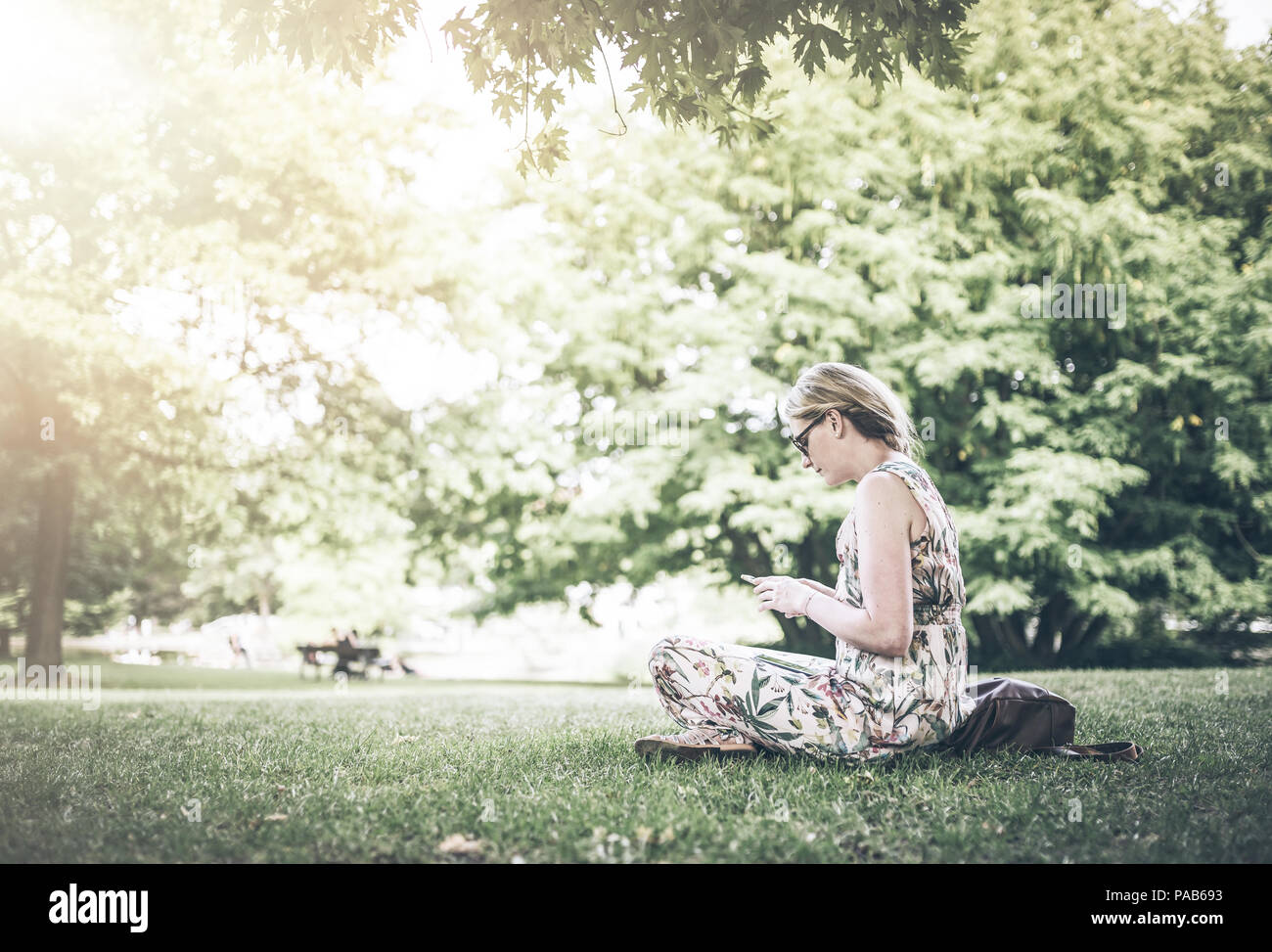 woman sitting cross-legged on lawn in public park using her smartphone on sunny summer day Stock Photo
