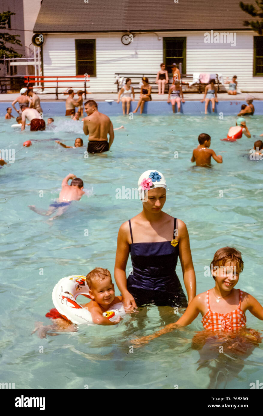 Woman wearing one piece bathing costume and a flowery swimming cap with young children in a busy swimming pool in Summer, Briarcliff Manor local pool, Westchester County, New York, USA in the 1960s Stock Photo