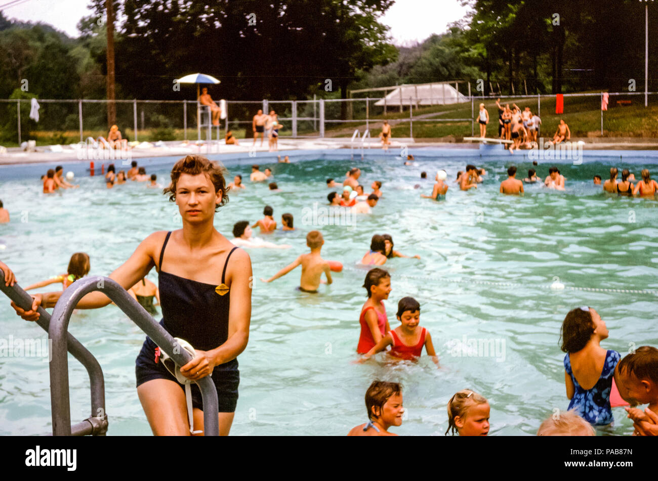 Woman wearing one piece bathing costume climbing a ladder out of a busy swimming pool, Briarcliff Manor local pool, Westchester County, New York, USA in 1960s Stock Photo