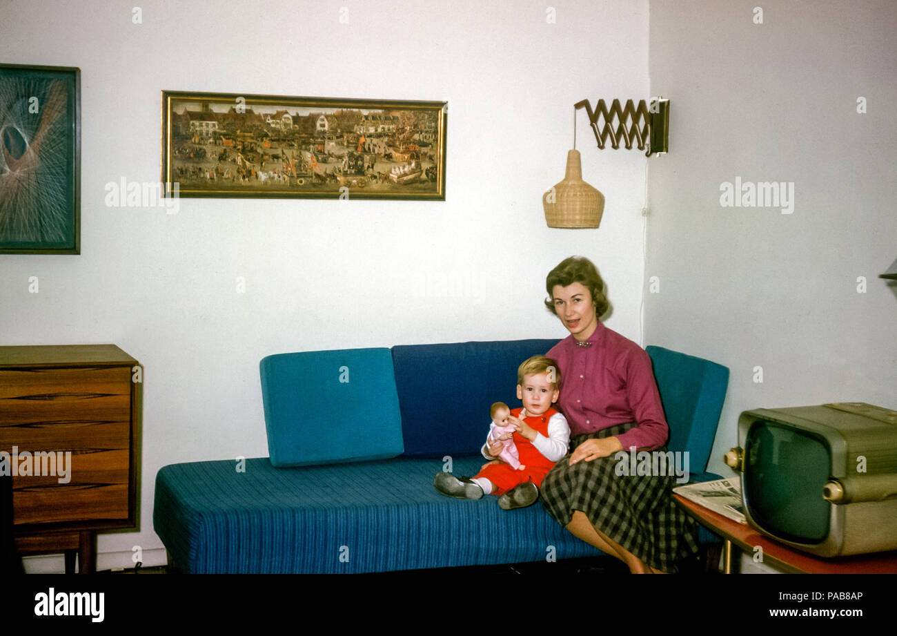 Young attractive woman or mother with toddler or two year old young boy holding a doll sitting on a day bed sofa in USA in the 1960s with an old fashioned television and The Triumph of Archduchess Isabella by Denys van Alsloot painting hanging on the wall Stock Photo