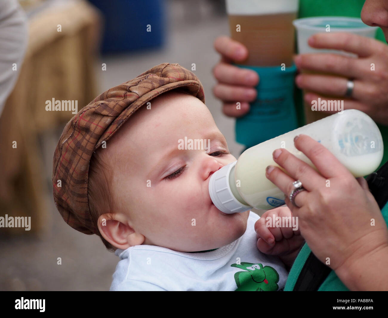 An adorable baby boy in a plaid hat is being bottle fed by Mom. Taken at the st. Patrick's Day Block Festival in Corpus Christi, Texas. Stock Photo