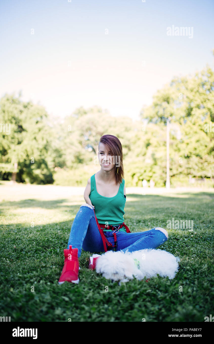 Portrait of beautiful smiling invalid young woman born without upper extremities enjoying with her little white puppy in park. Stock Photo