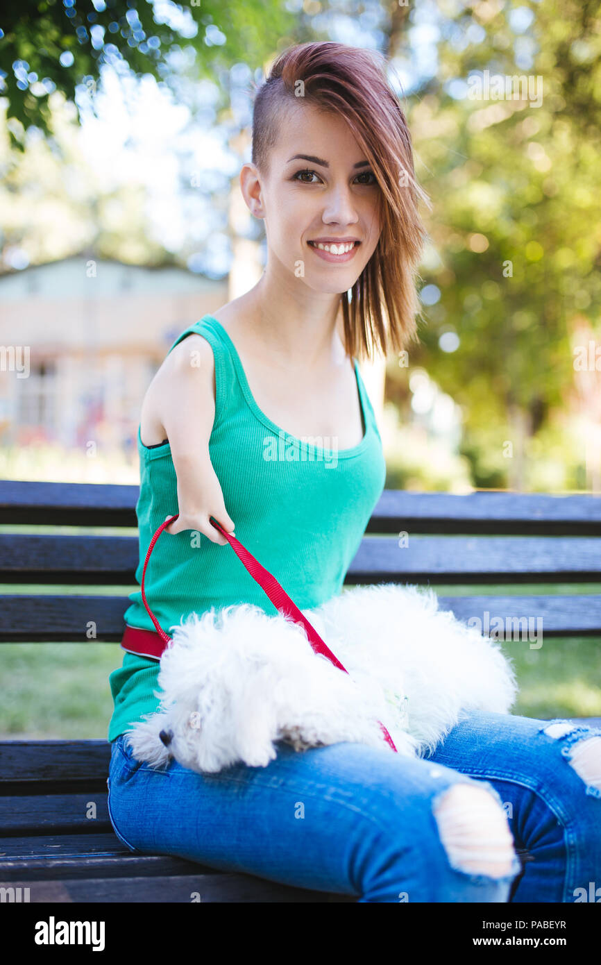 Portrait of beautiful smiling invalid young woman born without upper extremities holding her white little puppy and sitting on bench in park. Stock Photo
