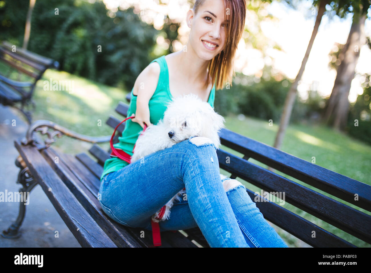 Portrait of beautiful smiling invalid young woman born without upper extremities holding her white little puppy and sitting on bench in park. Stock Photo