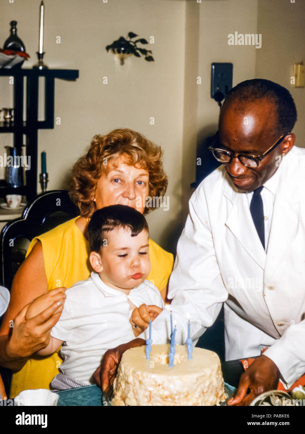 Child's birthday in the 1960s.  The 4 year old little boy is blowing out candles on a large birthday cake. He is sitting on his grandmother's lap as a black butler in a white jacket serves the birthday cake, USA Stock Photo