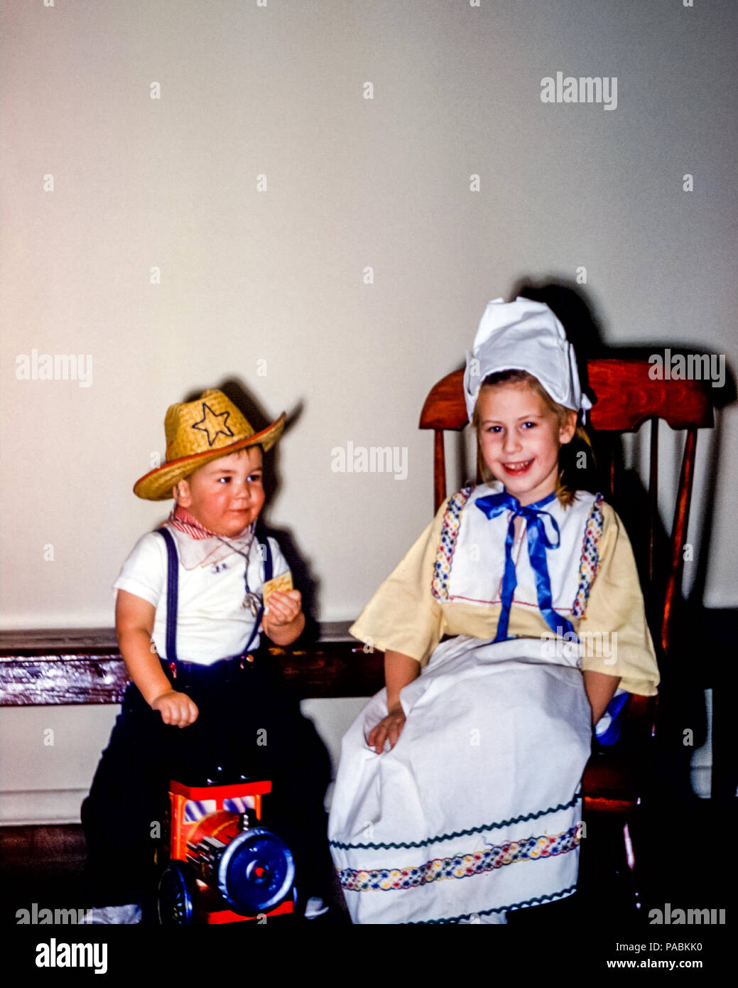 Young children in fancy dress costume for Halloween in USA in the 1950s.  The little four year old girl is wearing a traditional Dutch dress costume and the little boy is wearing a cowboy hat and kerchief sitting on a toy engine Stock Photo