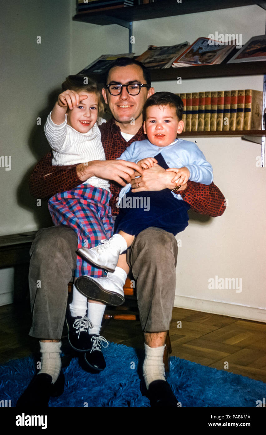 Family with father and two young children, a little girl and a boy toddler, sitting on his lap in a sitting room with book shelves with books and magazines in USA in the 1950s Stock Photo