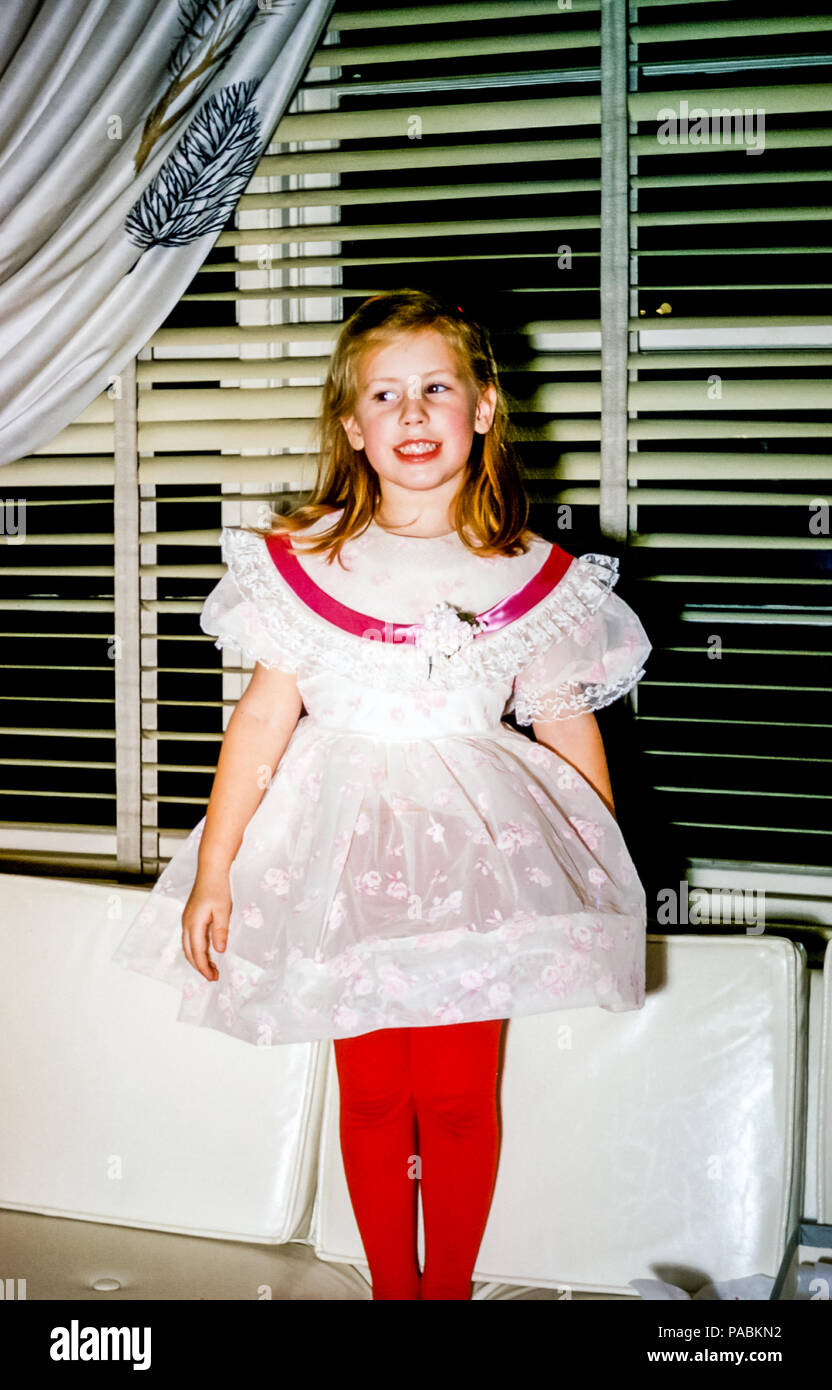 Four year old young girl with blonde hair wearing fancy party dress and red tights standing on a sofa, USA  in the 1950s Stock Photo