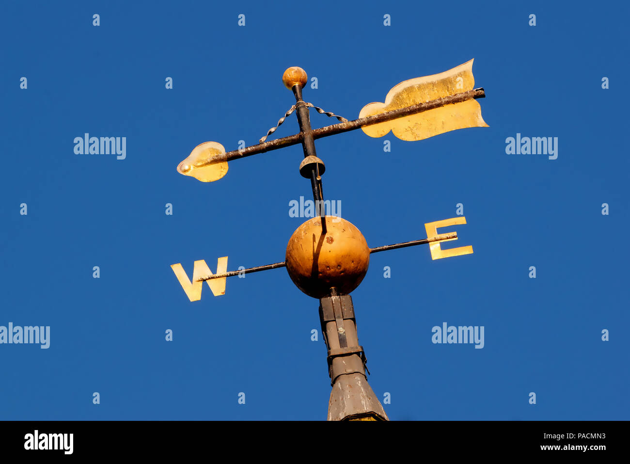 Gold coloured weather vane showing west and east with arrow against bright blue sky Stock Photo
