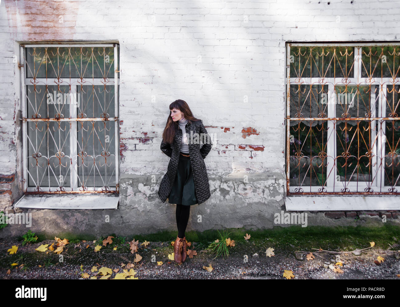 Beautiful sad girl stands against a white brick wall between two barred windows and looks aside Stock Photo