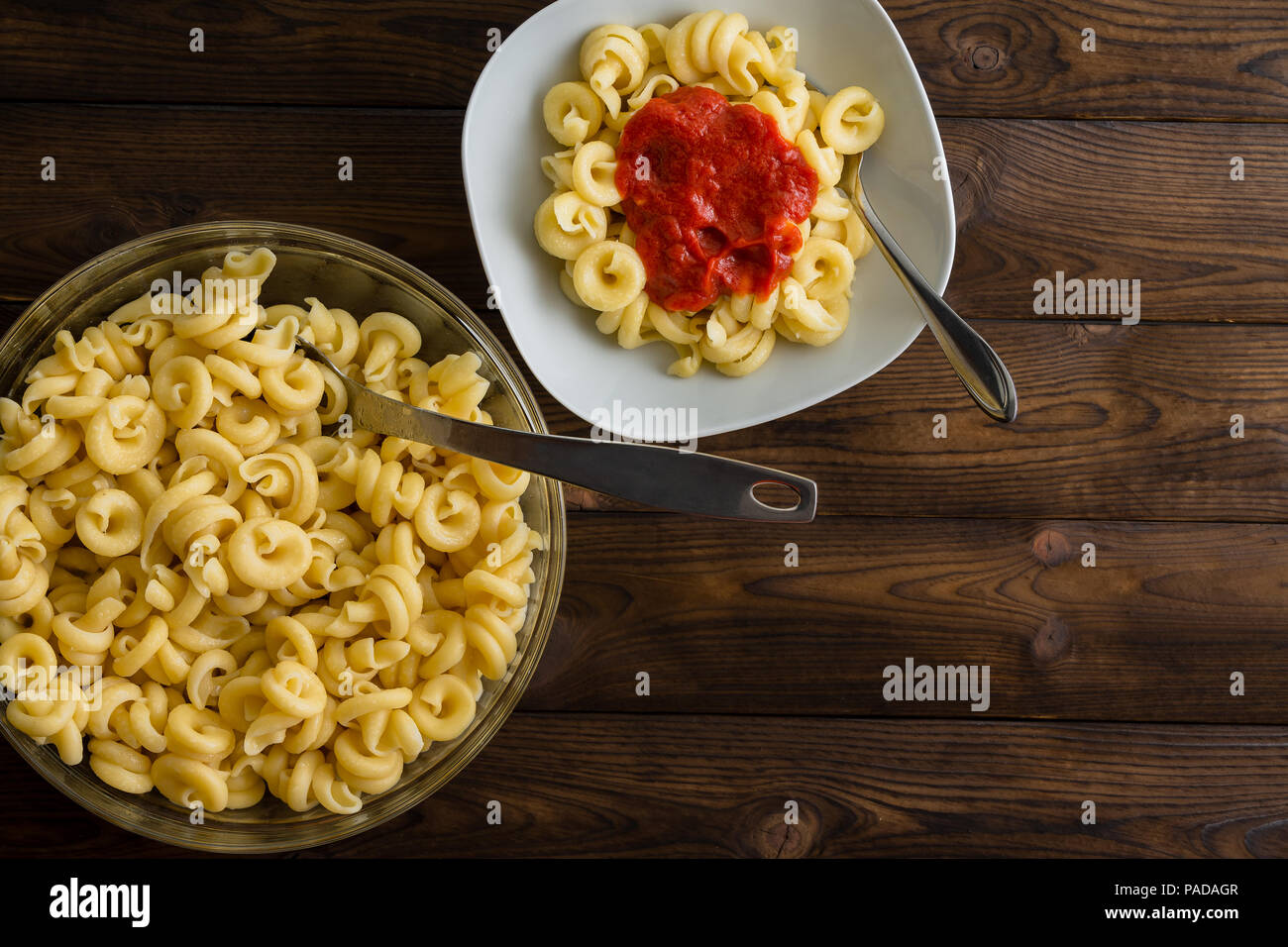 White square plate of pasta with tomato sauce next to clear glass dish of plain pasta over wood plank background. Includes copy space. Stock Photo