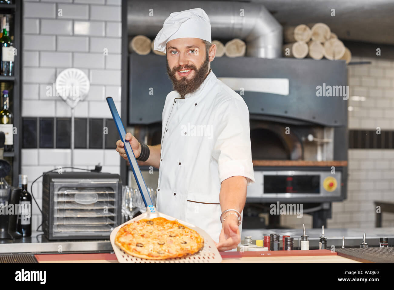 Front view of baker wearing chef's tunic and keeping pizza on metallic shovel. Working on spacy restaurant's kitchen with big oven and wooden timbers behind. Man looking at camera, feeling happy. Stock Photo