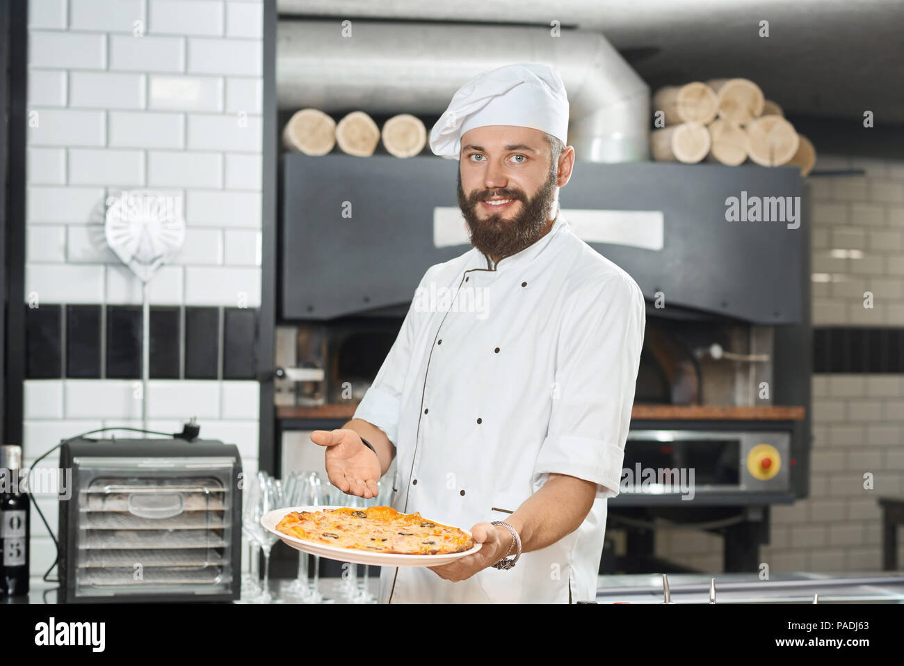 Smiling bearded pizzaiolo holding fresh baked mouthwatering pizza on big plate. Wearing white chef's tunic and a hat, working on restaurant kitchen with oven and alcohol bottles standing behind. Stock Photo