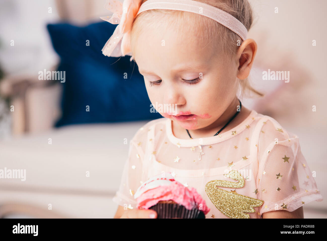 children's birthday. funny two-year-old Caucasian girl in pink dress standing to bedroom of house the background of couch and eating, holding cake des Stock Photo