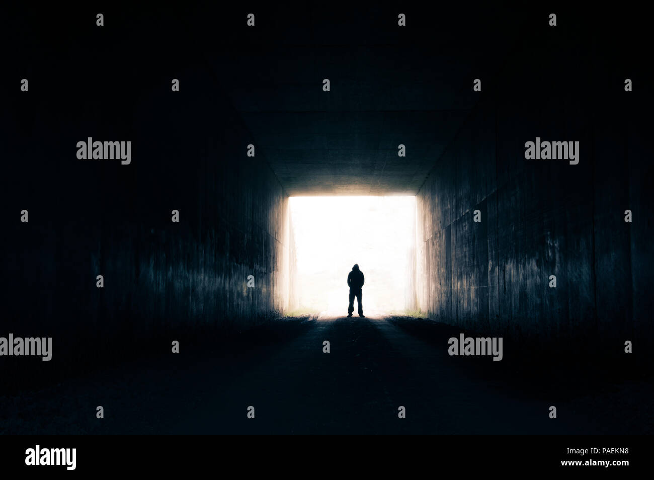 A silhouette of a man standing at the end of a tunnel looking out on bright white light Stock Photo