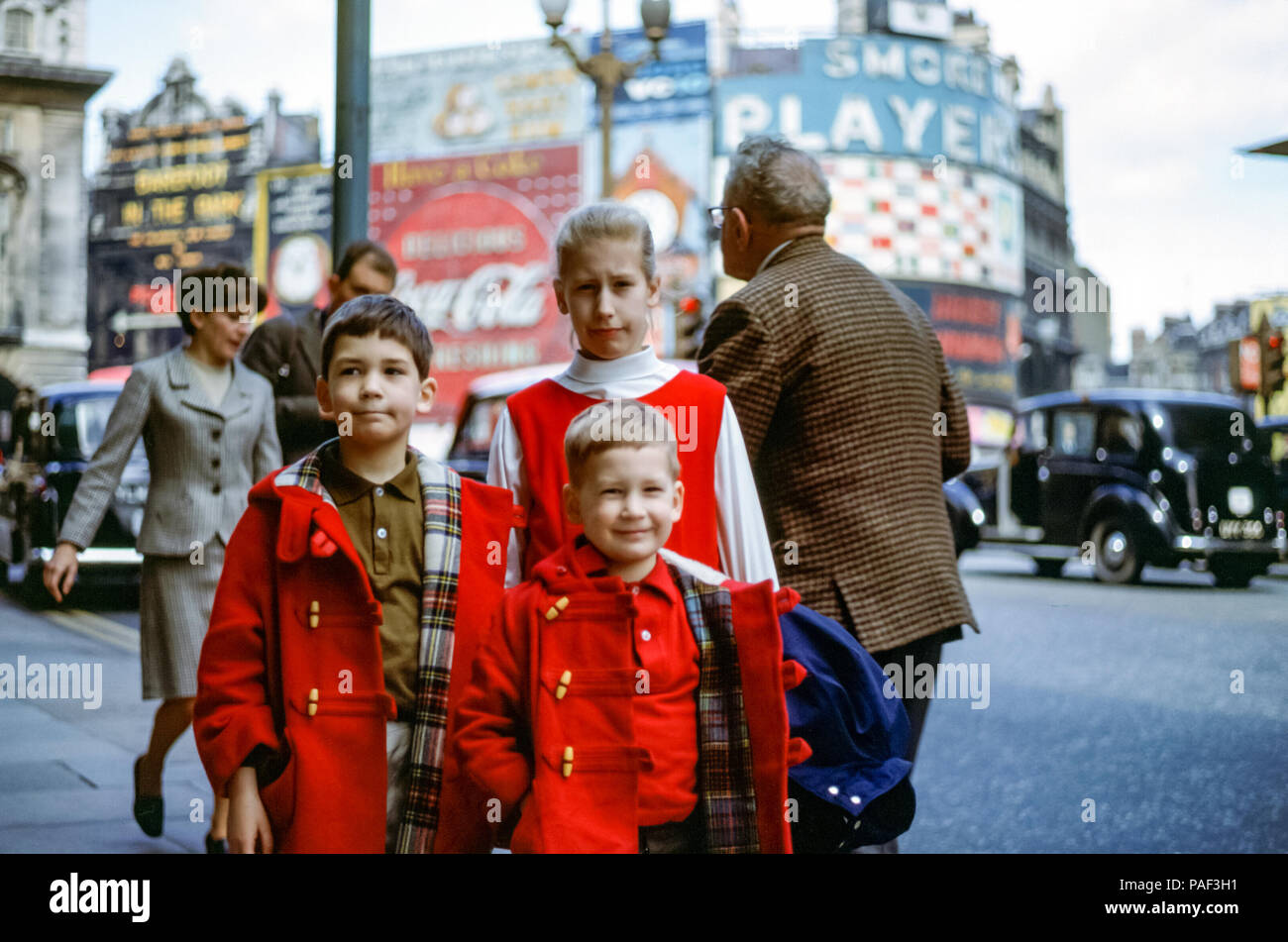 Three children in a family portrait, two boys and a girl wearing red in Piccadilly Circus, London, England in the 1960s. The boys are wearing matching red duffel coats. Piccadilly Theatre advertises the play Barefoot in the Park with neon advertisements for Coca Cola and Players cigarettes Stock Photo