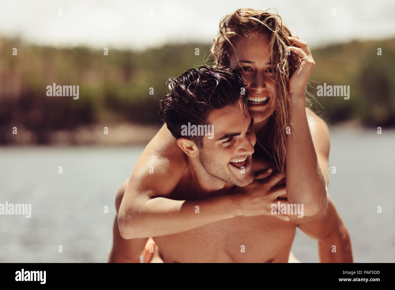 Portrait of happy young man piggybacking his girlfriend. Cheerful young couple enjoying their vacation on lakeside. Stock Photo