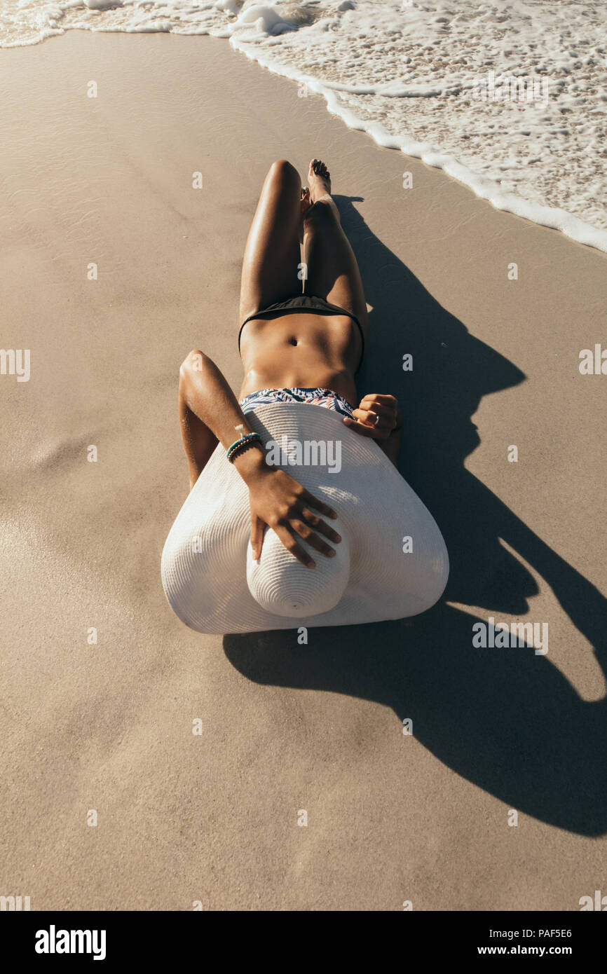Woman in bikini sunbathing lying near the seashore wearing sun hat with sea waves reaching her. Woman on vacation wearing sun hat relaxing at the beac Stock Photo