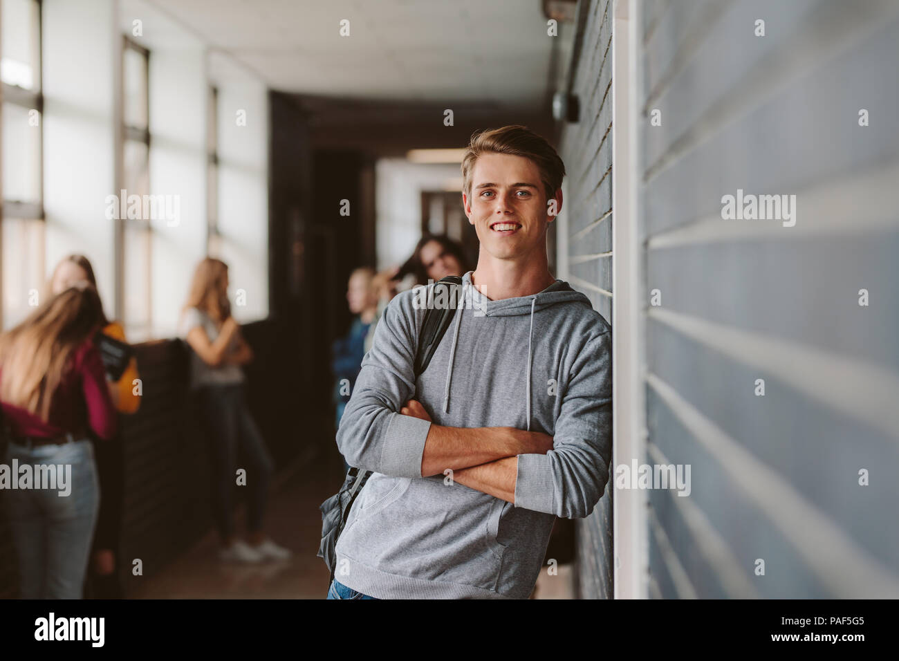 Portrait of handsome young university student standing and leaning to a wall in college corridor with other students at the back. Caucasian male stude Stock Photo
