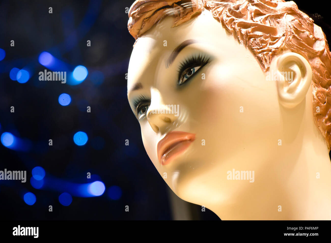 Close up of one female mannequin doll with short curly ginger hair displayed n the shop window with blurred blue lights in the background , profile vi Stock Photo