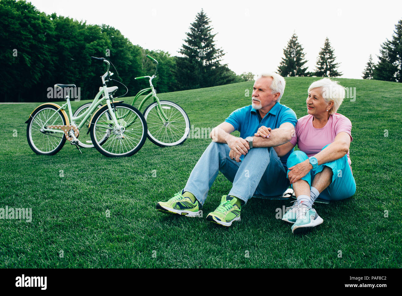 positive old couple embracing, sits on the grass, with their bicycles Stock Photo