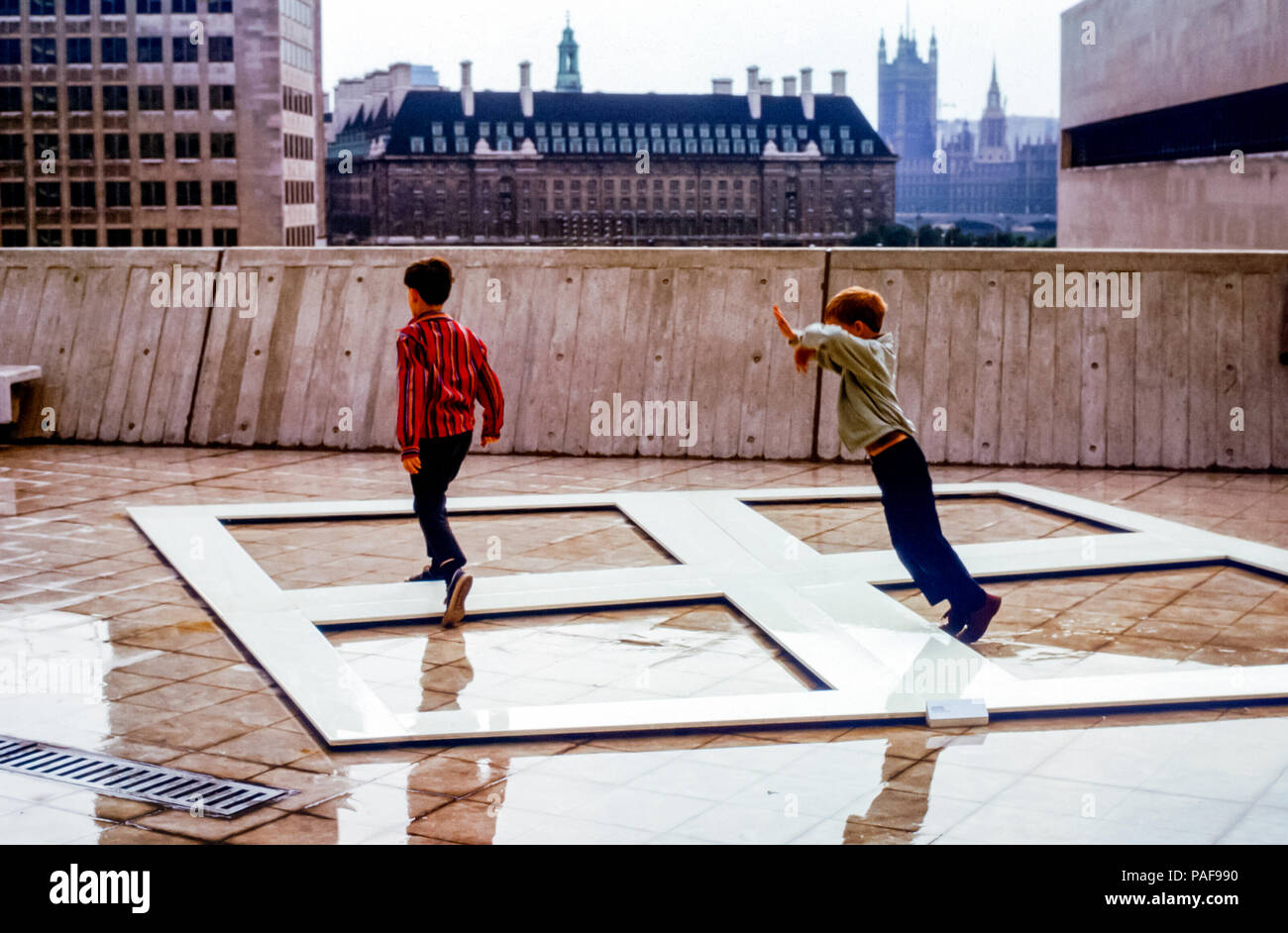 Young boys having fun jumping over modern artwork on the outdoor deck of Hayward Gallery, South Bank, London, England, UK in the 1960s. The small boy is about to trip and fall on the edge of the art installation Stock Photo