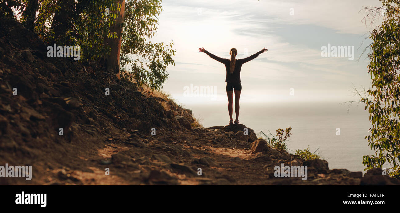 Rear view of woman standing on mountain looking at view with arms wide open. Female runner with arms outstretched on mountain peak. Stock Photo