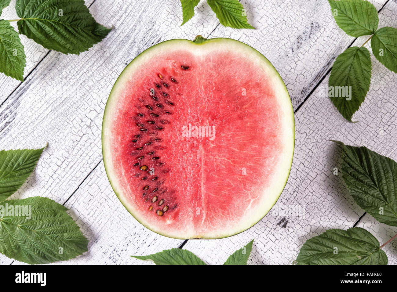 Half of ripe red watermelon partly with and partly without seeds surrounded by green leaves on white old wooden table with cracks. Healthy eating and  Stock Photo