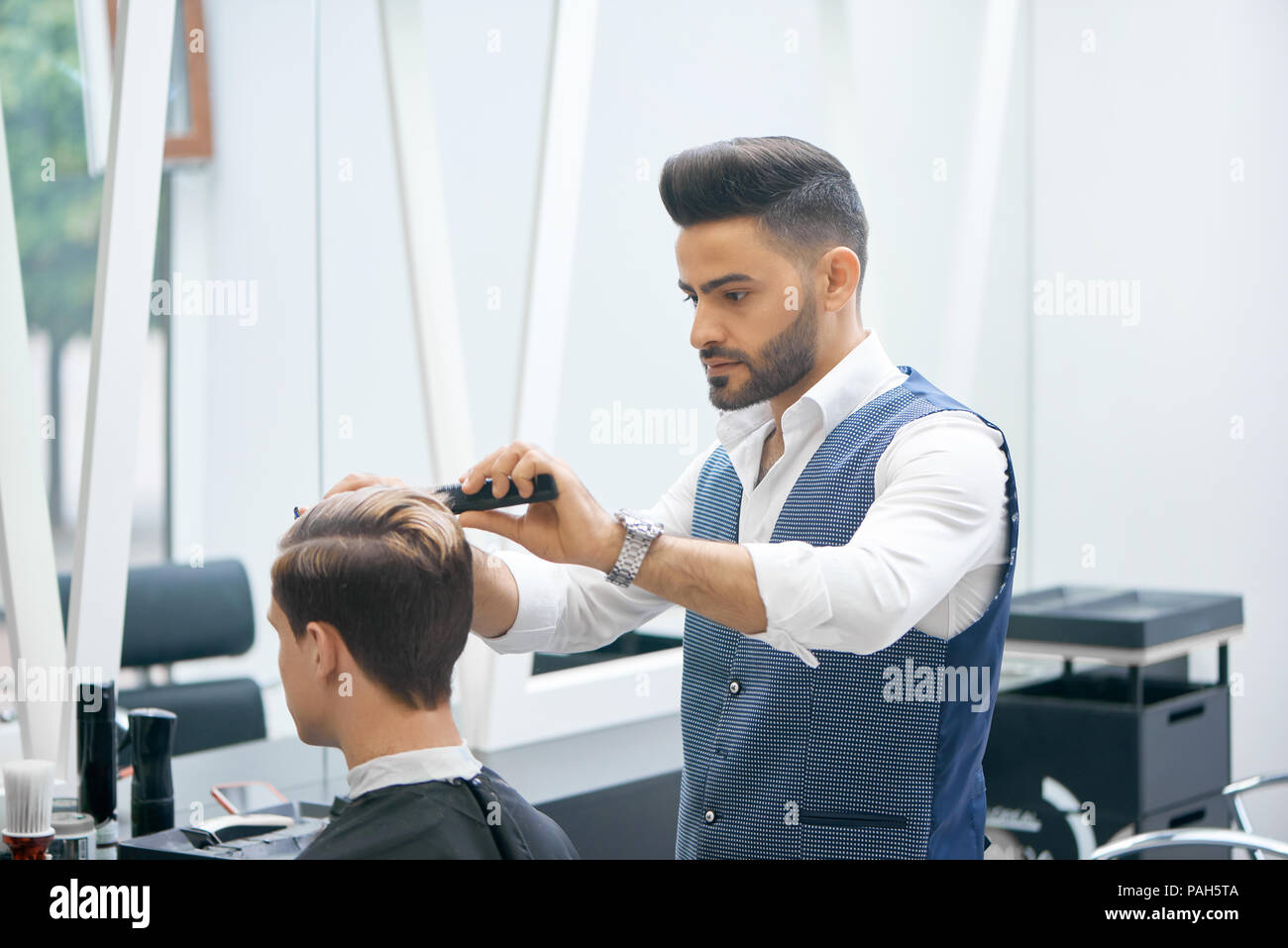 Barber doing new haircut for young client using black plastic comb. Wearing white casual shirt, grey waistcoat, watch. Looking concentrated, loving his job. Model covered with special black cape. Stock Photo