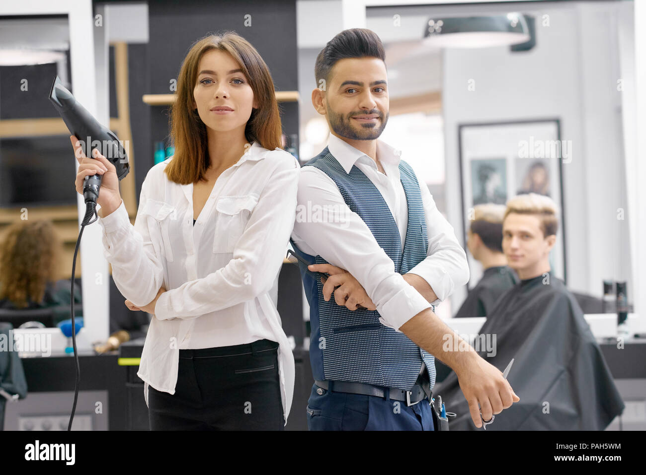 Two hairstylers posing standing in modern beaty salon, young client sitting behind. Hairdressers