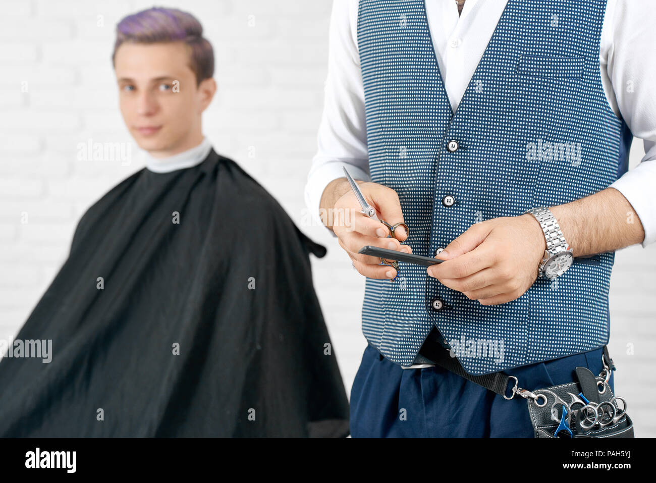 Cropped photo of barber keeping black plastic comb and scissors istanding in front ofyoung client. Boy having toned in violet color hair and looking at camera. Master wearing scissors case, waistcoat. Stock Photo