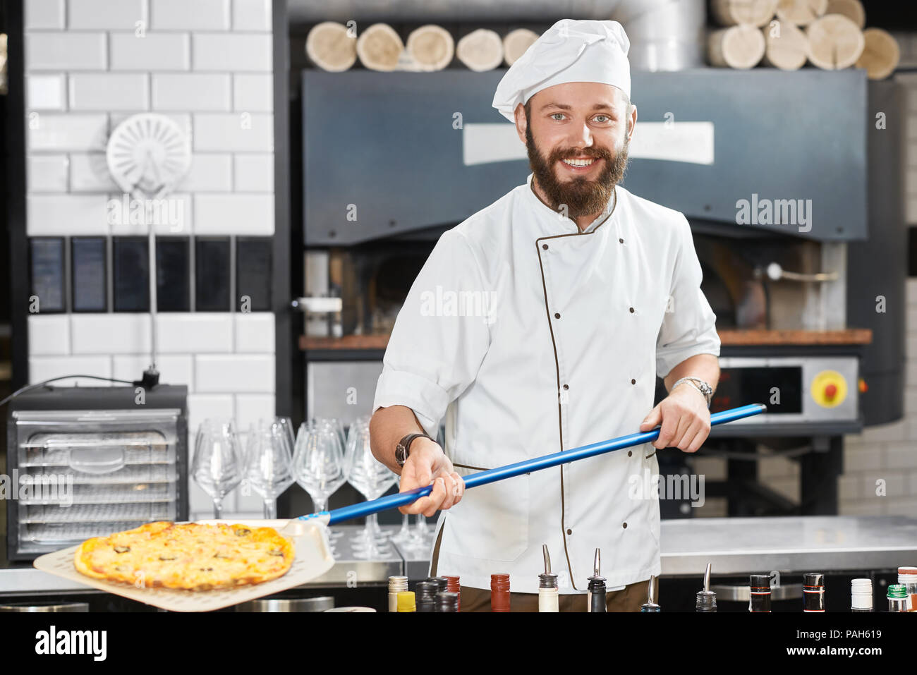 Smiling, happy baker keeping pizza on long blue metallic shovel, looking at camera. Wearing white chef's tunic and a cap,looking at camera. Working in restaurant's kitchen with alcohol shelves nearby. Stock Photo