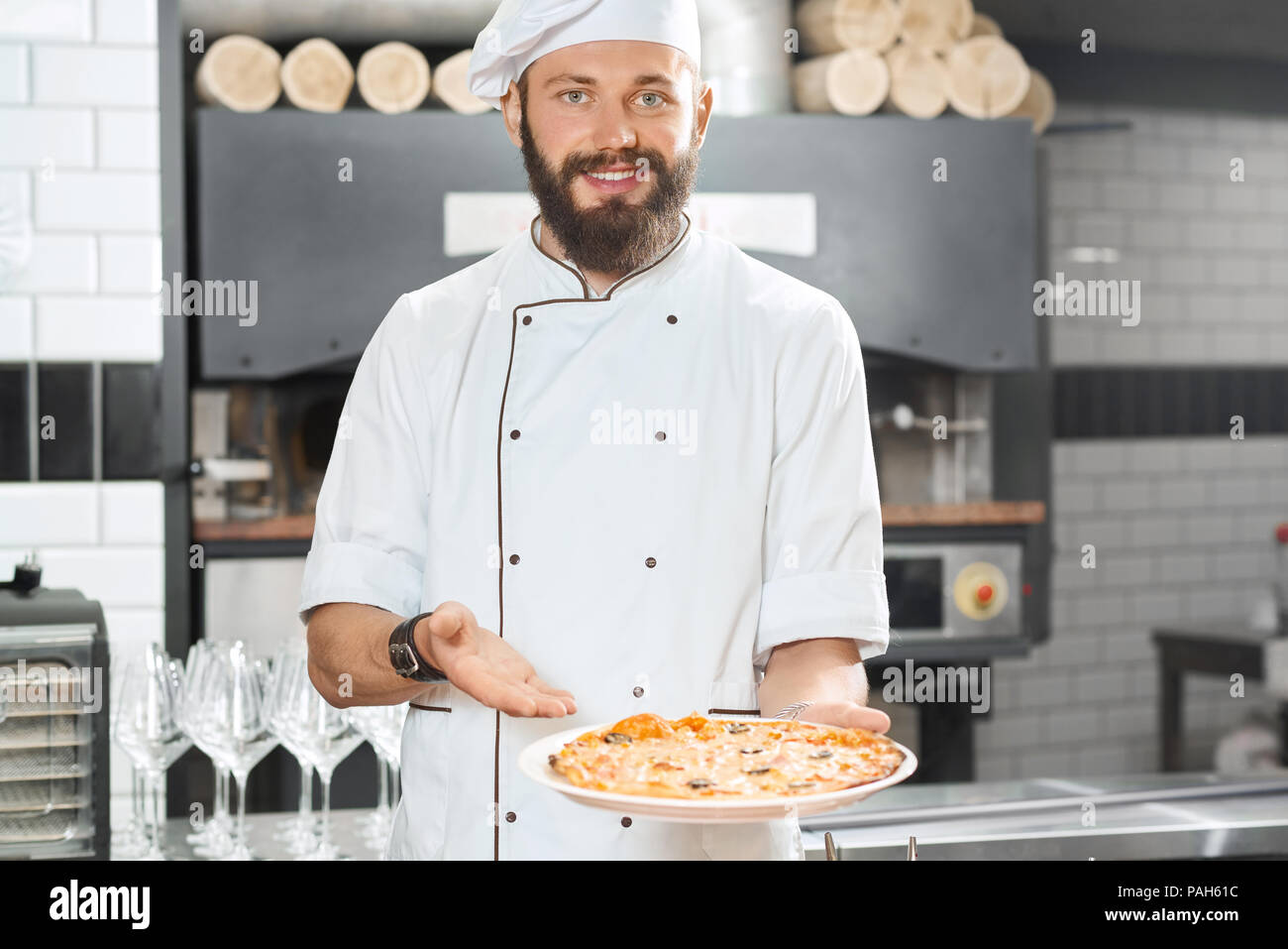 Smiling pizzaiolo holding fresh baked mouthwatering, delicious pizza. Wearing white chef's tunic working on restaurant kitchen with oven, wooden timbers, wine glasses set standing on background. Stock Photo