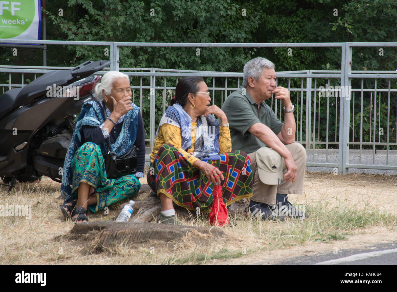 Three older Nepalese Gurkha people sitting on a treestump watching the Farnborough International Airshow, 2018 during the heatwave with parched grass Stock Photo