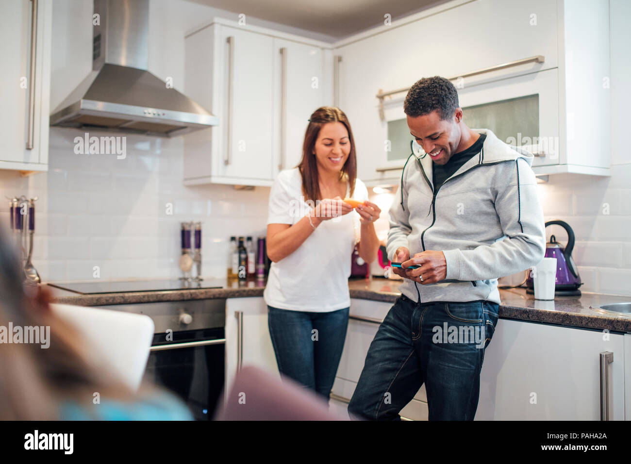 Mid adult couple are relaxing in the kitchen of their home together. The woman is eating a slice of orange and the man is using his smart phone. Stock Photo
